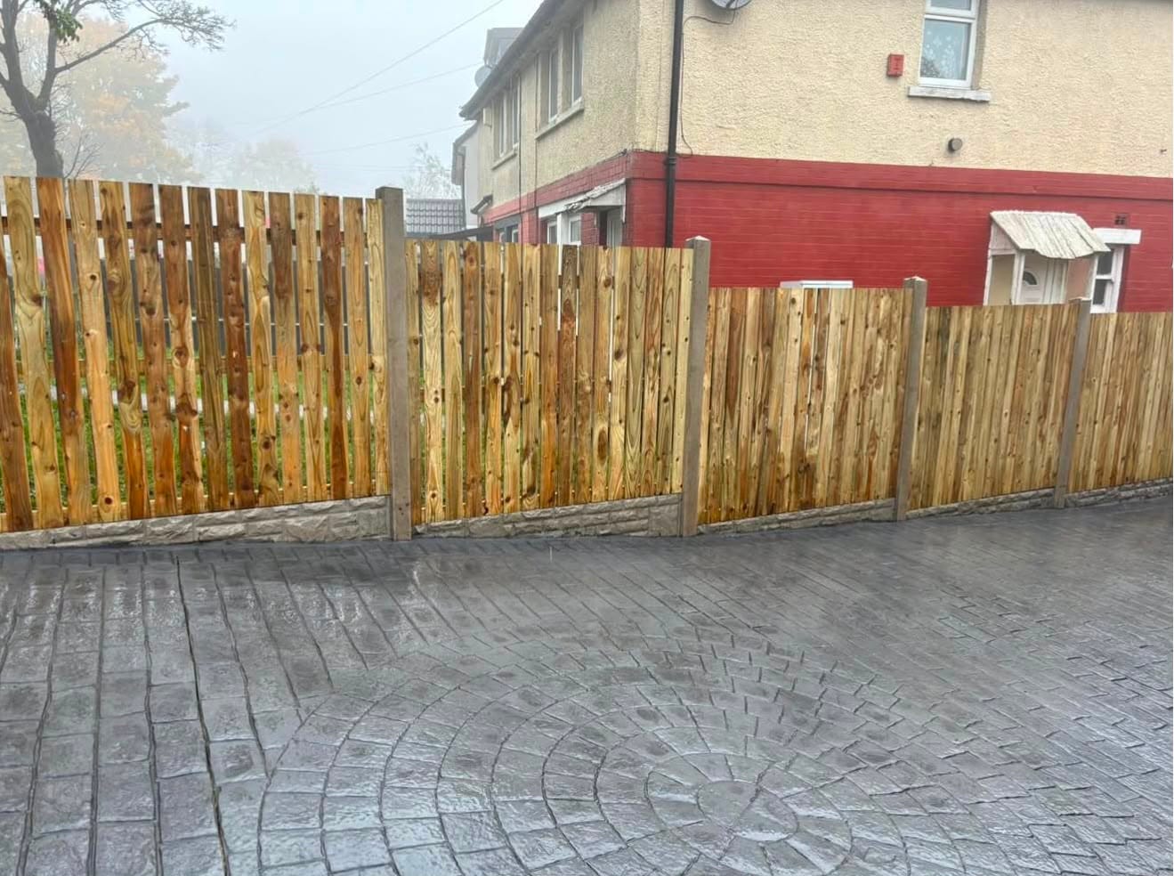 Grey imprinted concrete driveway with a cobble circle feature beside new timber fencing on a natural stone plinth on a misty morning