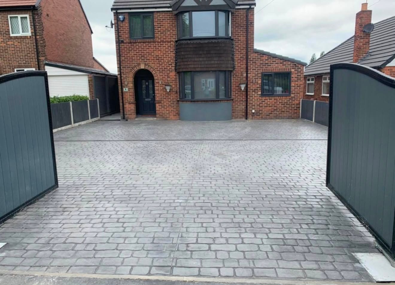 Wide grey cobble-sett imprinted concrete driveway between black timber gates at a red-brick detached house
