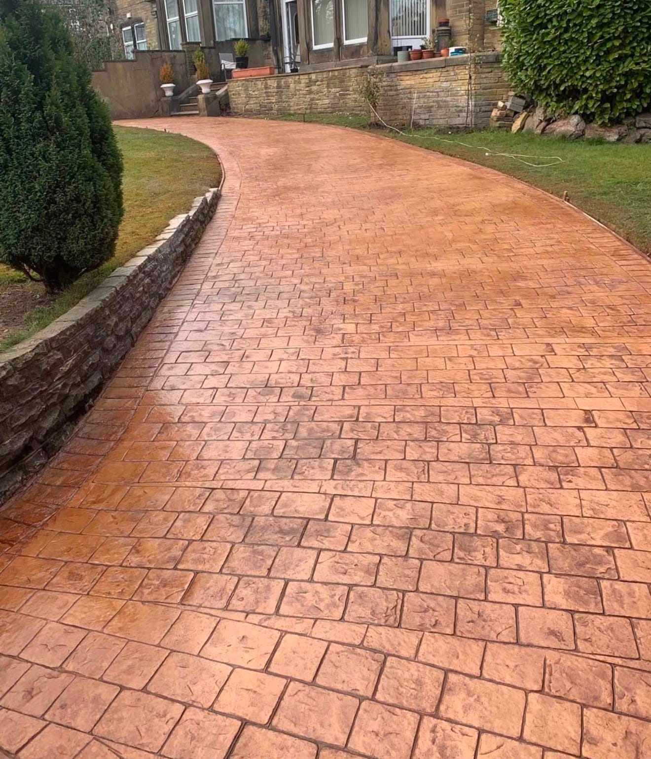 Curving warm brindle imprinted concrete driveway sweeping past a natural stone retaining wall and lawn to a period stone house
