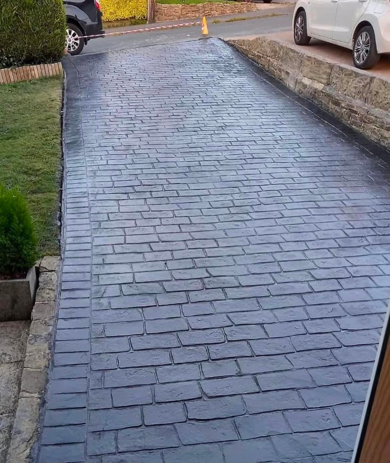 Slate-grey brick-pattern imprinted concrete driveway edged against a natural stone garden wall, with a traffic cone at the top